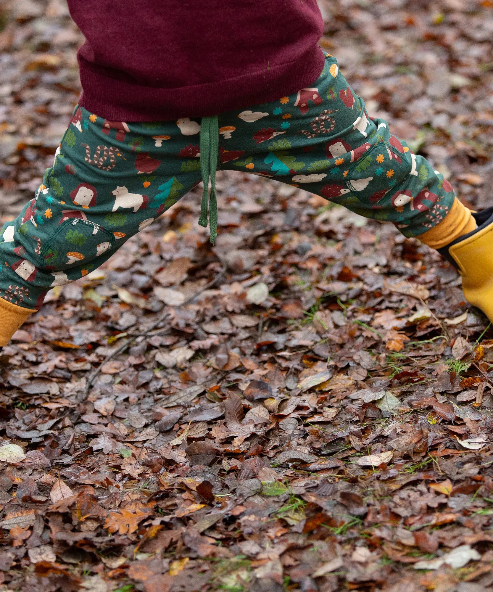 Child wearing the Little Green Radicals cosy organic cotton knee patch joggers with a woodland design print with yellow gold wellington boots.