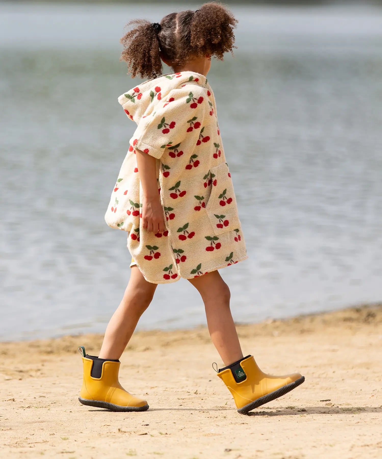 Child wearing the Little Green Radicals little cherries cream organic cotton kids hooded towel poncho.The poncho has a popper closure on the neck and a kangaroo pocket on the front. 