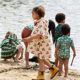 Side view of a child wearing the Little Green Radicals little cherries cream organic cotton kids hooded towel poncho.The poncho has a popper closure on the neck and a kangaroo pocket on the front. 