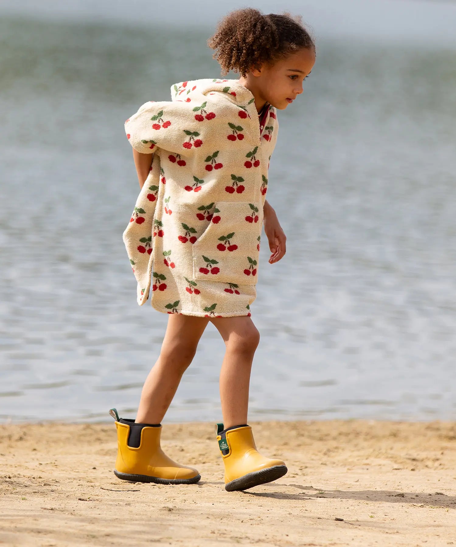 Child wearing the Little Green Radicals little cherries cream organic cotton kids hooded towel poncho with yellow wellington boots. The poncho has a popper closure on the neck and a kangaroo pocket on the front. 
