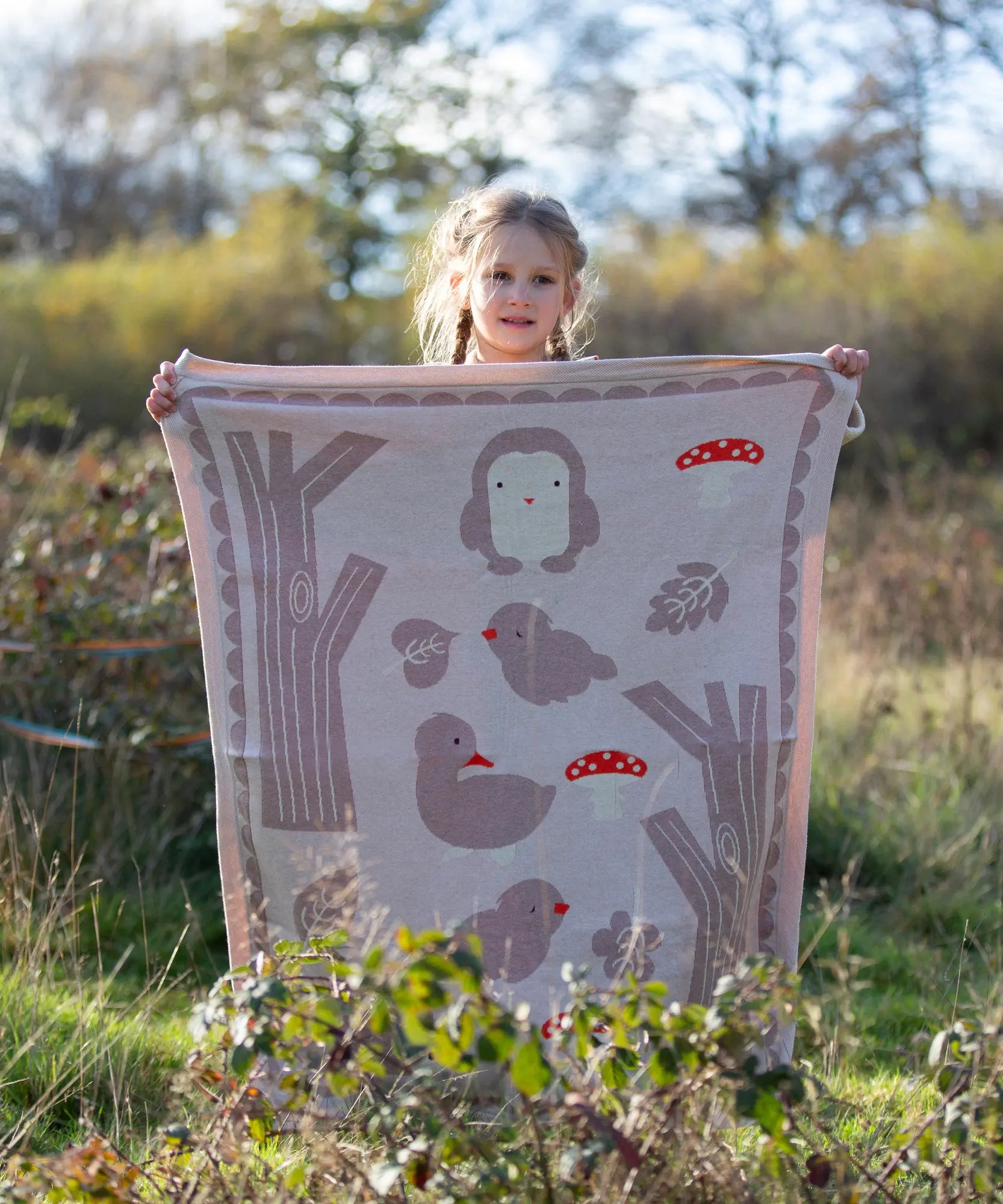 Child standing behind a Little Green Radicals cream folk tales knitted organic cotton baby blanket that they are holding up in front of them.