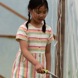 A child wearing the Little Green Radicals cream rainbow striped short sleeve button front dress. The dress has side pockets.