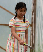 A child wearing the Little Green Radicals cream rainbow striped short sleeve button front dress. The dress has side pockets.
