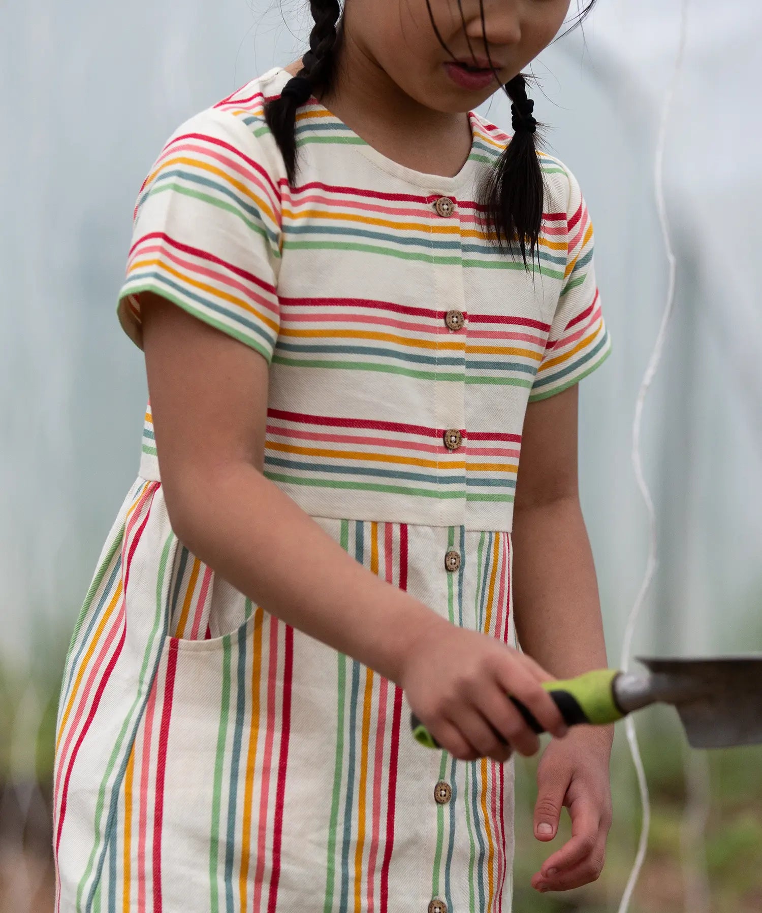 A close up of the button up front and side pockets on the Little Green Radicals children's cream rainbow striped short sleeve dress being worn by a child.