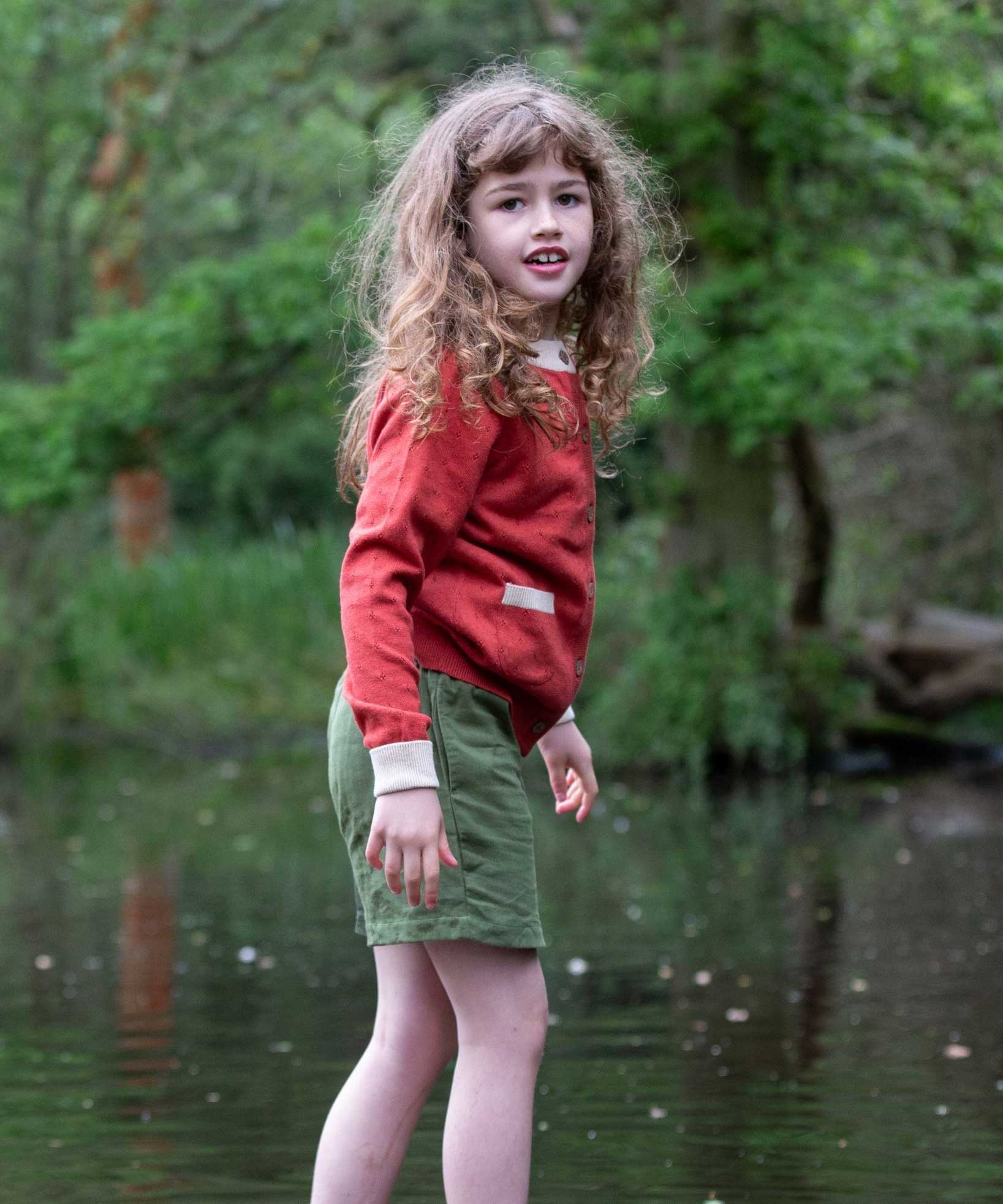 A child wearing a Little Green Radicals From One To Another Burnt Ochre Pointelle Knitted Cardigan with green coloured shorts. Little green Radicals have a wide range of organic cotton, Fairtrade children's clothing and outerwear available here at Babipur.