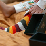 Child wearing the rainbow striped socks from the Little Green Radicals folk tales ankle socks 3 pack. Open green gift box placed next to the child's feet.