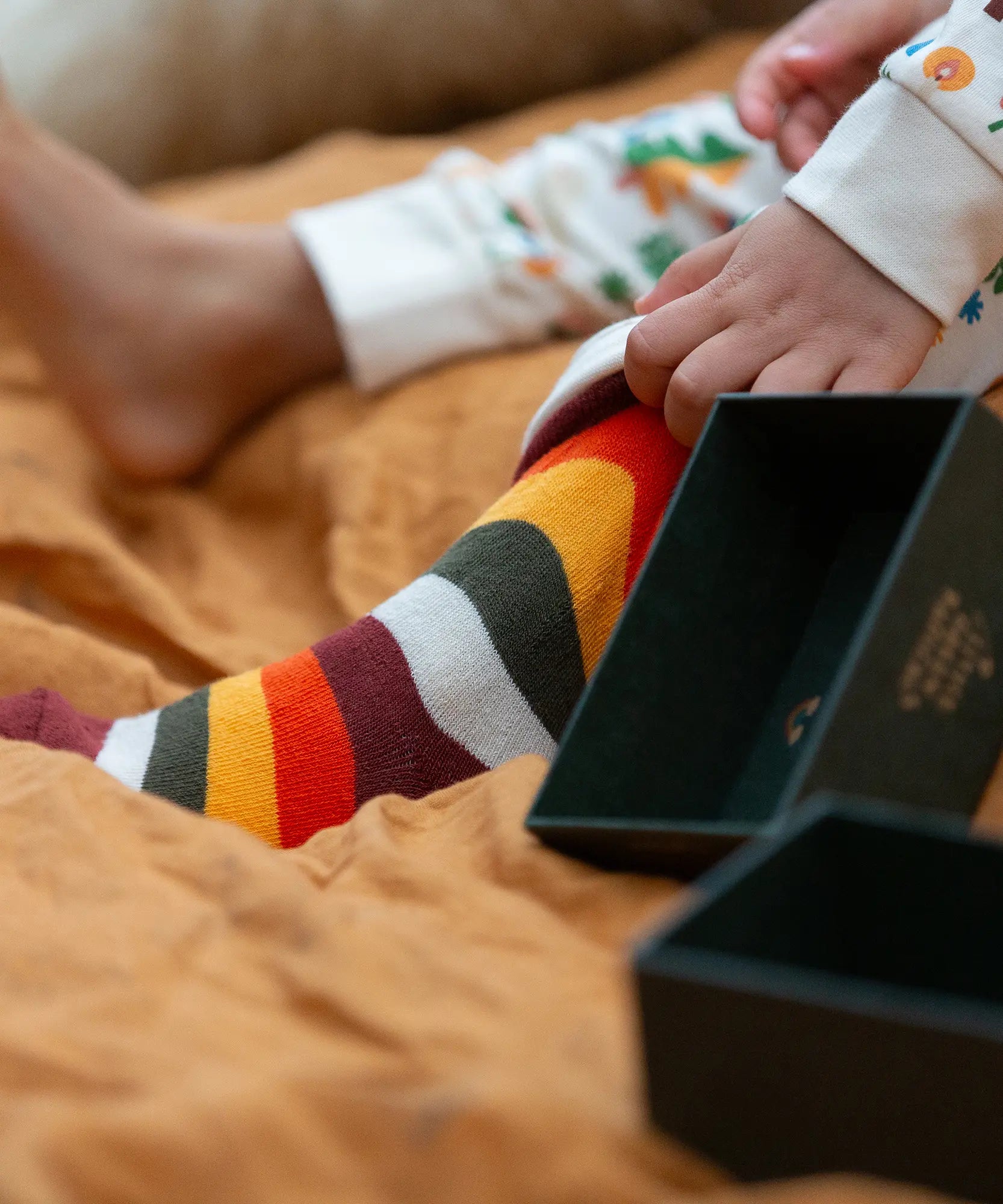 Child wearing the rainbow striped socks from the Little Green Radicals folk tales ankle socks 3 pack. Open green gift box placed next to the child's feet.