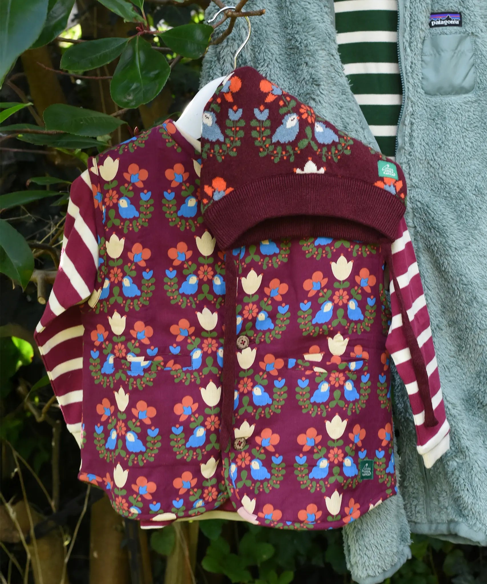 Little Green Radicals Organic Sherpa Reversible Gilet featuring the Folk Birds print with a knitted hat featuring the same print with a long sleeved striped top shown underneath.