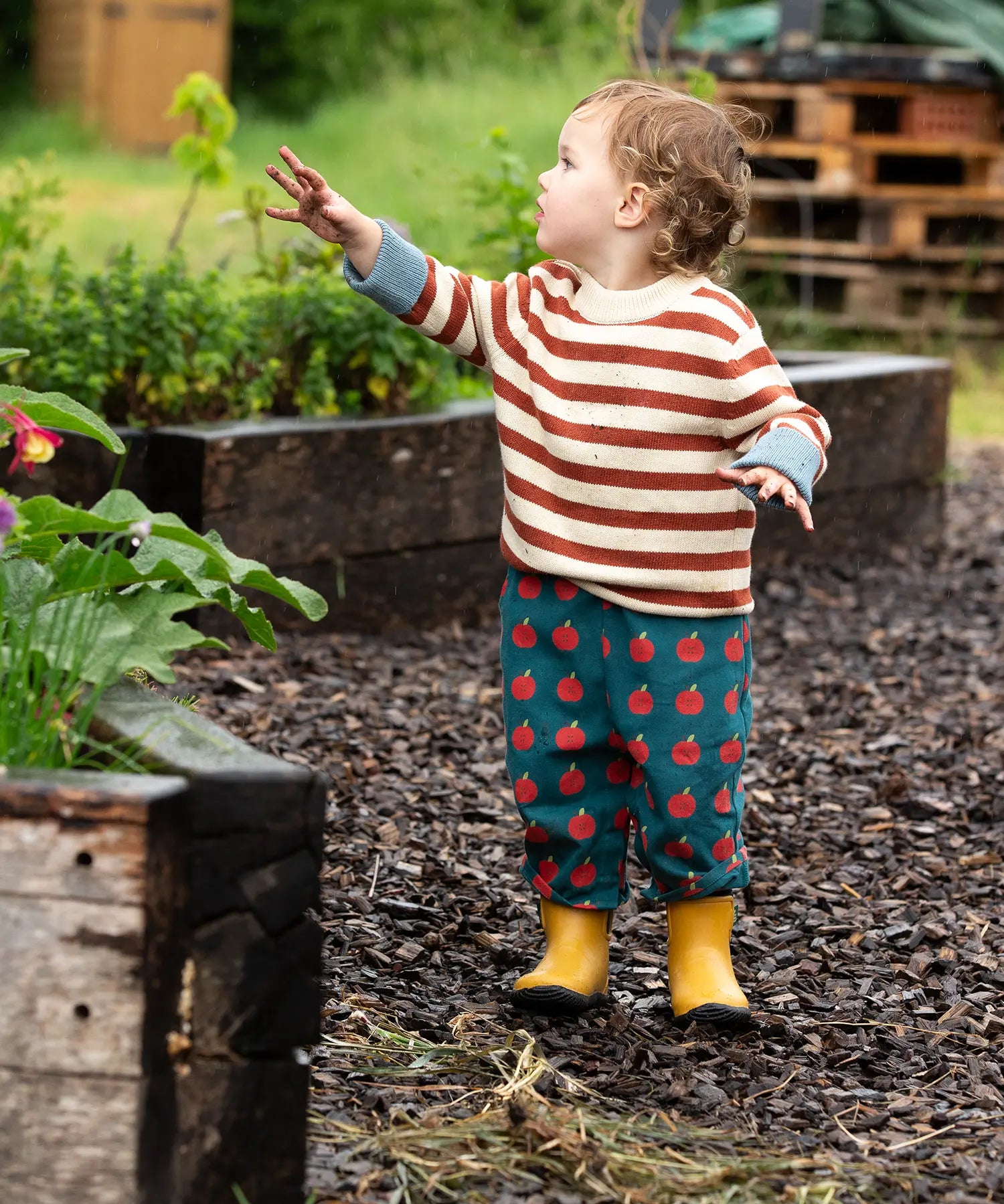 Young child wearing the Little Green Radicals ginger striped knitted jumper with repeat apple print trousers and yellow gold wellington boots.
