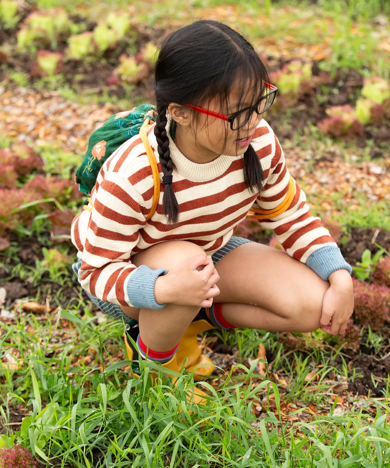 A child kneeling down wearing the Little Green Radicals ginger striped knitted jumper with blue and white pin stripe shorts and yellow gold wellington boots.