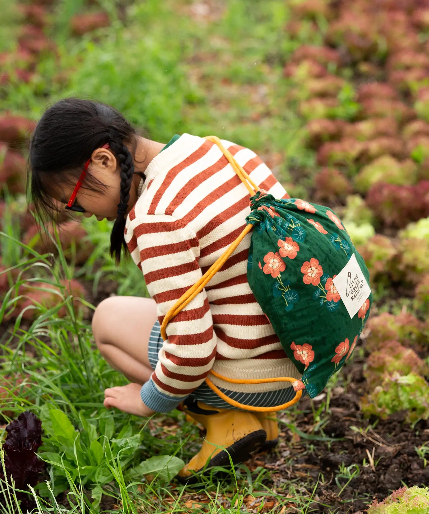 A child kneeling down wearing the Little Green Radicals ginger striped knitted jumper with blue and white pin stripe shorts and yellow gold wellington boots.