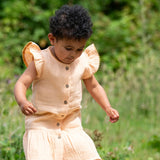 Child wearing the Little Green Radicals coral pink dropped waist children's dress with ochre yellow pom pom ribbon edging on the sleeves and collar.
