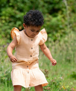 Child wearing the Little Green Radicals coral pink dropped waist children's dress with ochre yellow pom pom ribbon edging on the sleeves and collar.