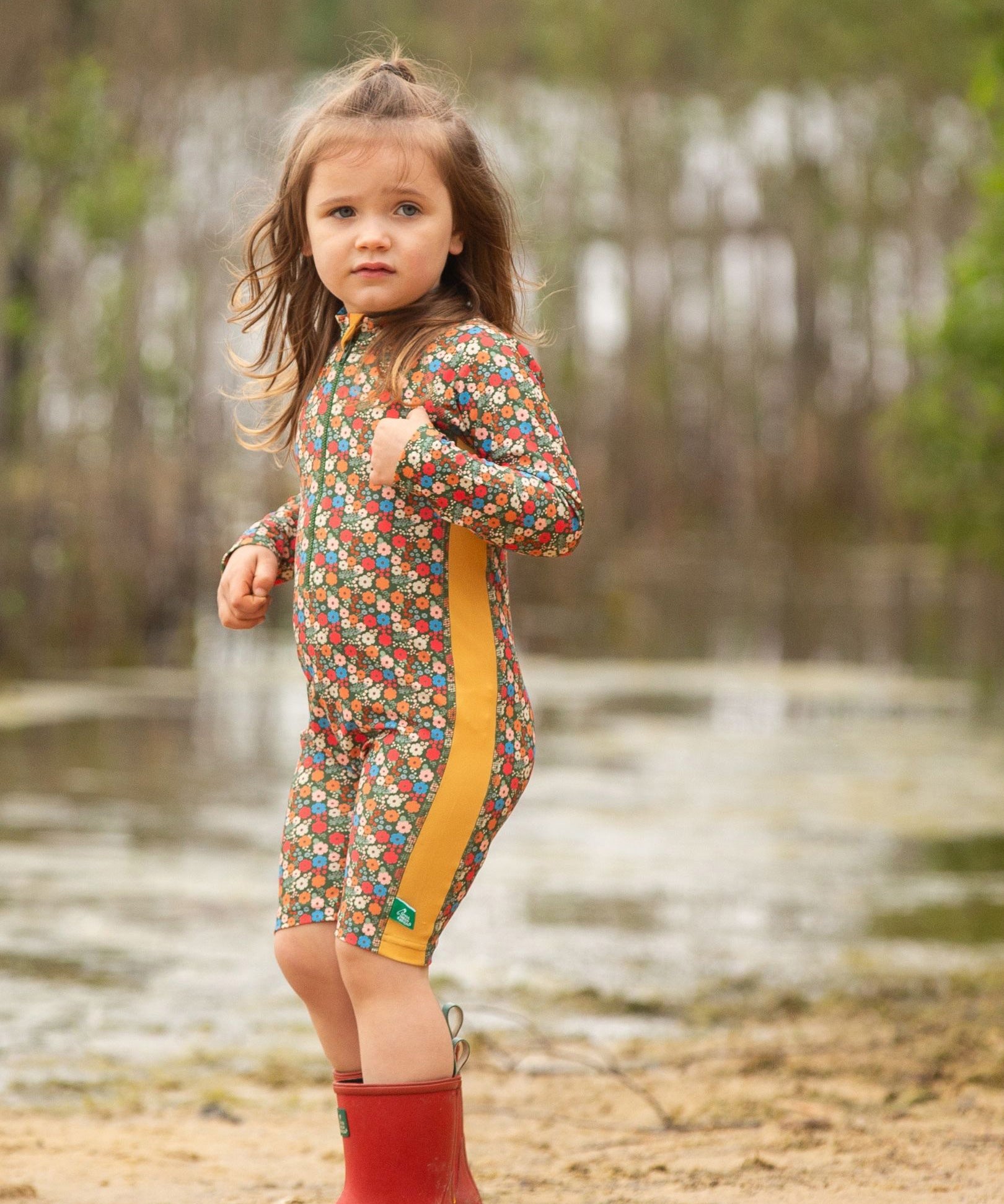 A child wearing the Little Green Radicals Vintage Flowers UPF 50+ Recycled Sunsafe Sunsuit with a pair of red wellington boots.
