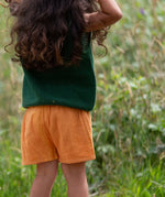Back of a child wearing the Little Green Radicals golden ochre kids shorts organic cotton shorts with a dark green knitted top.