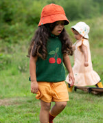 Child wearing the Little Green Radicals golden ochre kids shorts organic cotton shorts with a dark green knitted top and orange bucket hat.