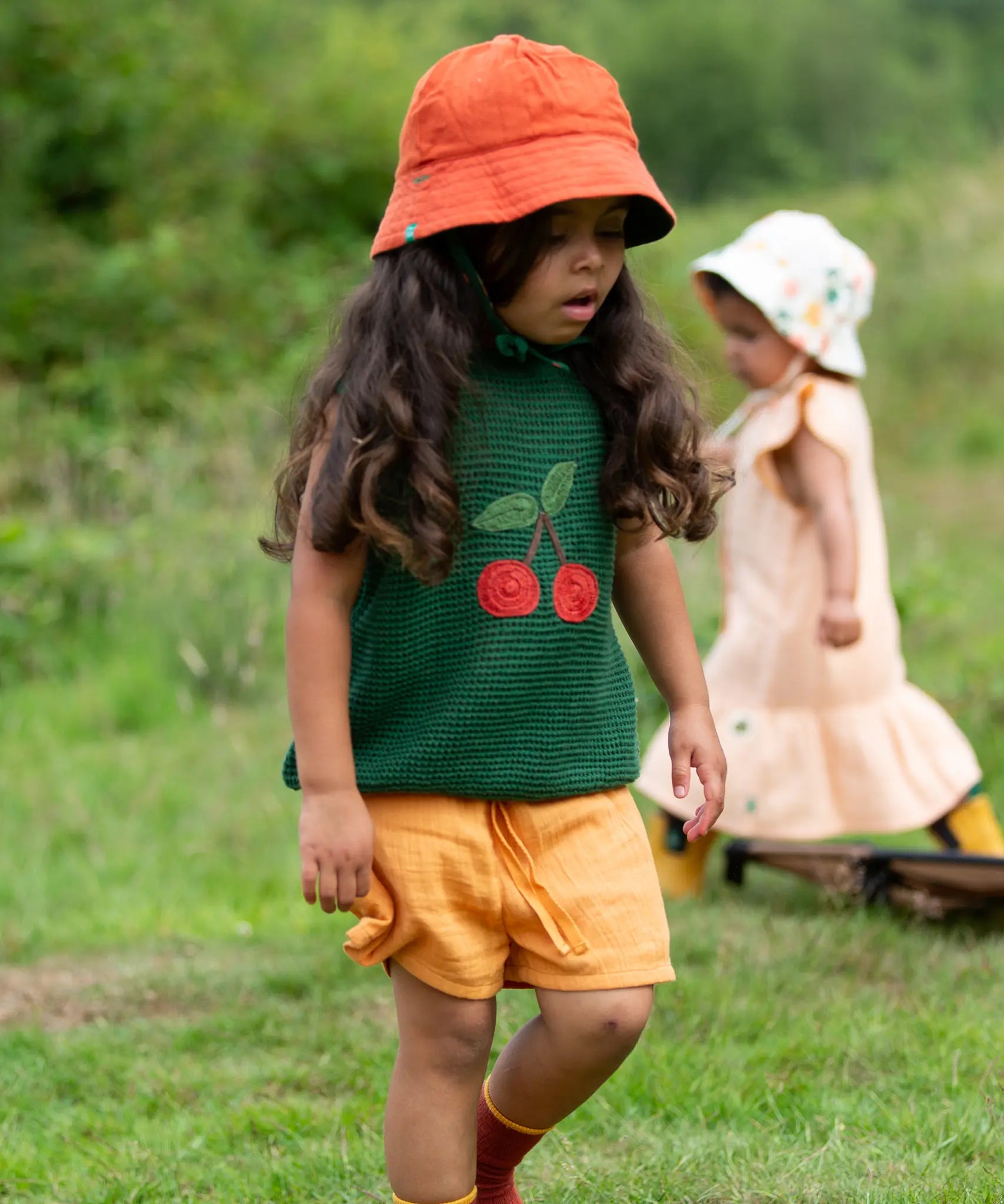 Child wearing the Little Green Radicals golden ochre kids shorts organic cotton shorts with a dark green knitted top and orange bucket hat.