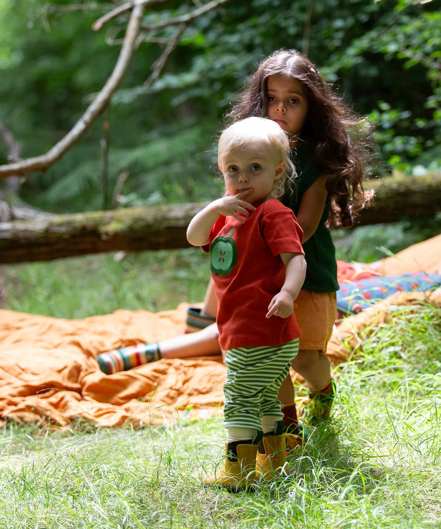 Child wearing the Little Green Radicals green apple applique organic short sleeve red t-shirt with cherry knee patch joggers and yellow wellington boots.