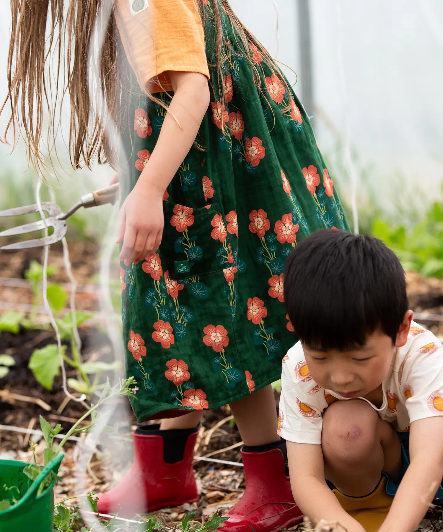 Close up of the side pockets on the Little Green Radicals green orange blooms repeat print reversible pinny kids dress being worn with a yellow t-short underneath and red wellington boots.