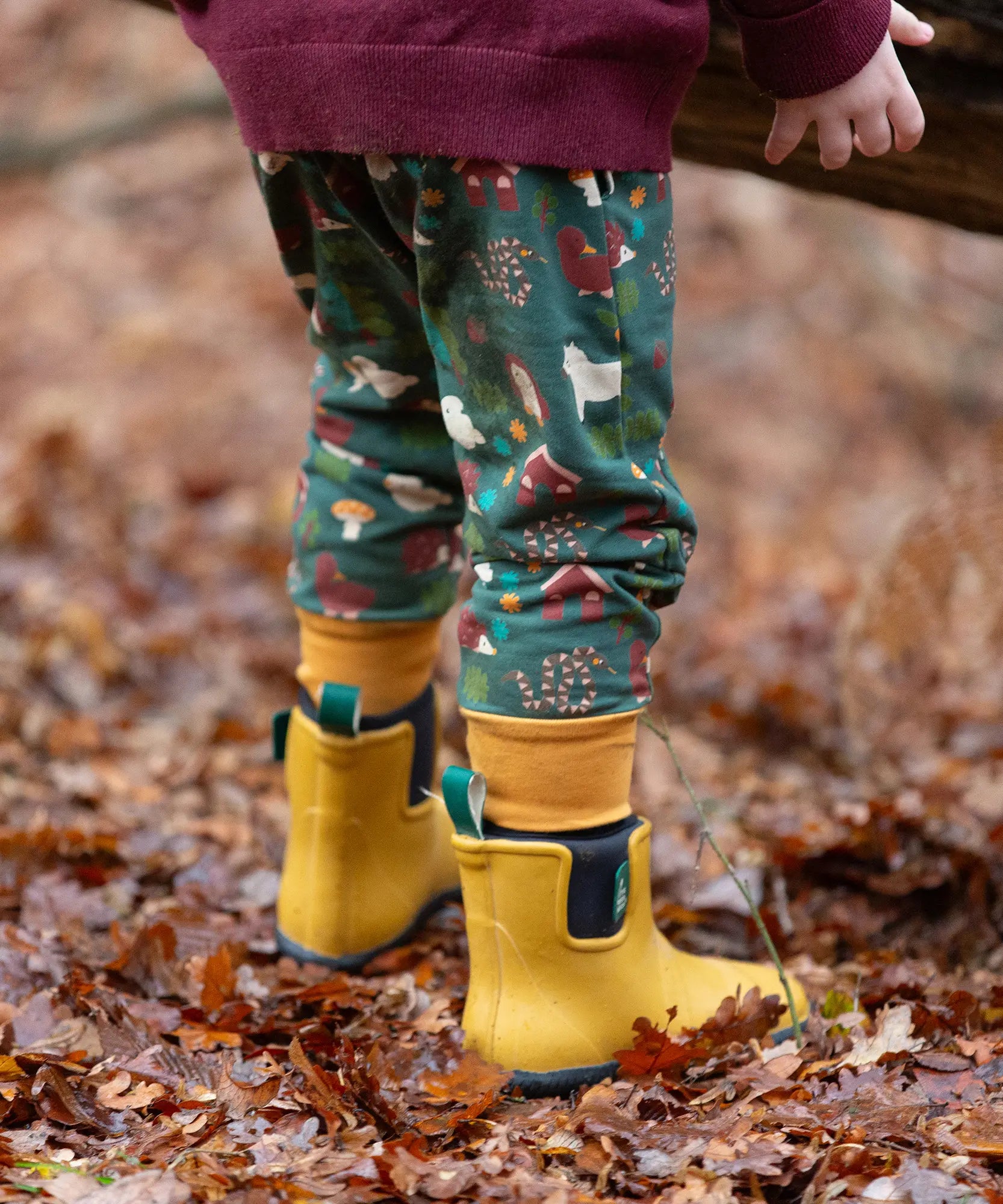Child wearing the Little Green Radicals cosy organic cotton knee patch joggers with a woodland design print with yellow gold wellington boots.