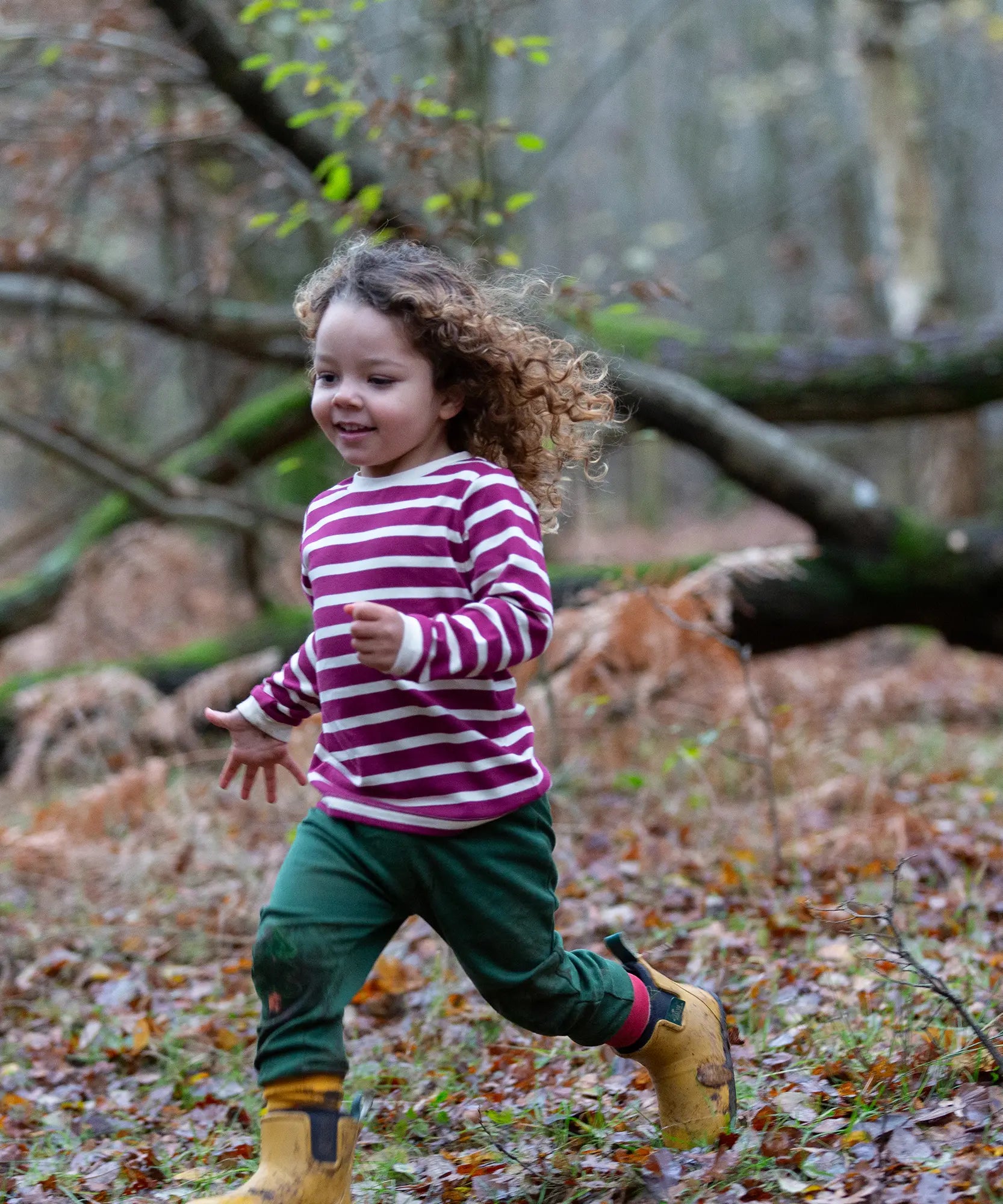 Child wearing the Little Green Radicals berry purple striped long sleeve top with green joggers and yellow gold wellington boots.