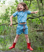 A child wearing the Little Green Radicals Kids Organic Cotton Blue Clouds Loose Fit Short Sleeve Henley Top with a pair of striped blue shorts. The child is jumping and has a pair of red wellies on.