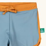 Close up of the Little Green Radicals hazy blue kids swim shorts with gold waist band and tie and binding. A small green logo tag can be seen on the side.