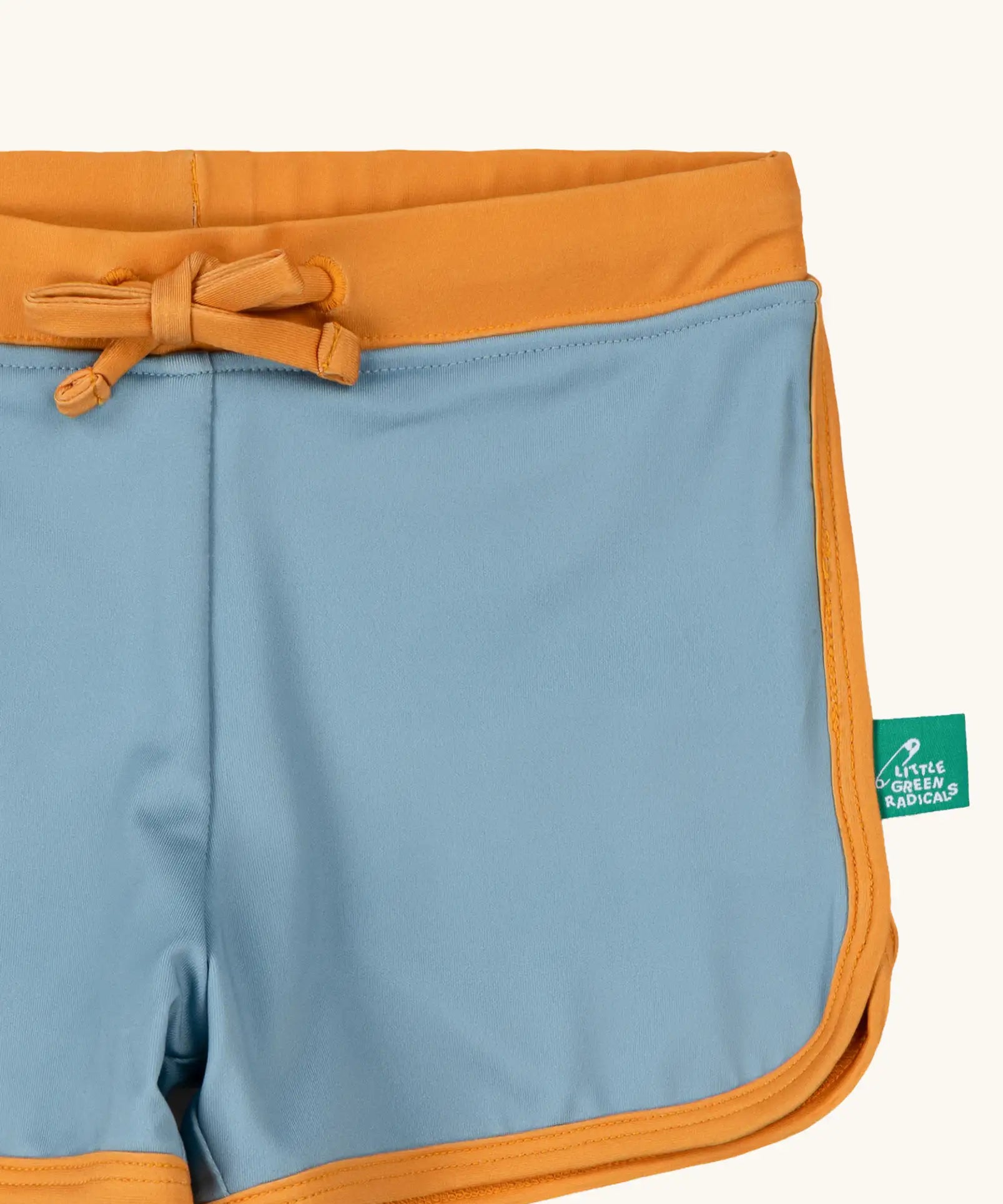 Close up of the Little Green Radicals hazy blue kids swim shorts with gold waist band and tie and binding. A small green logo tag can be seen on the side.