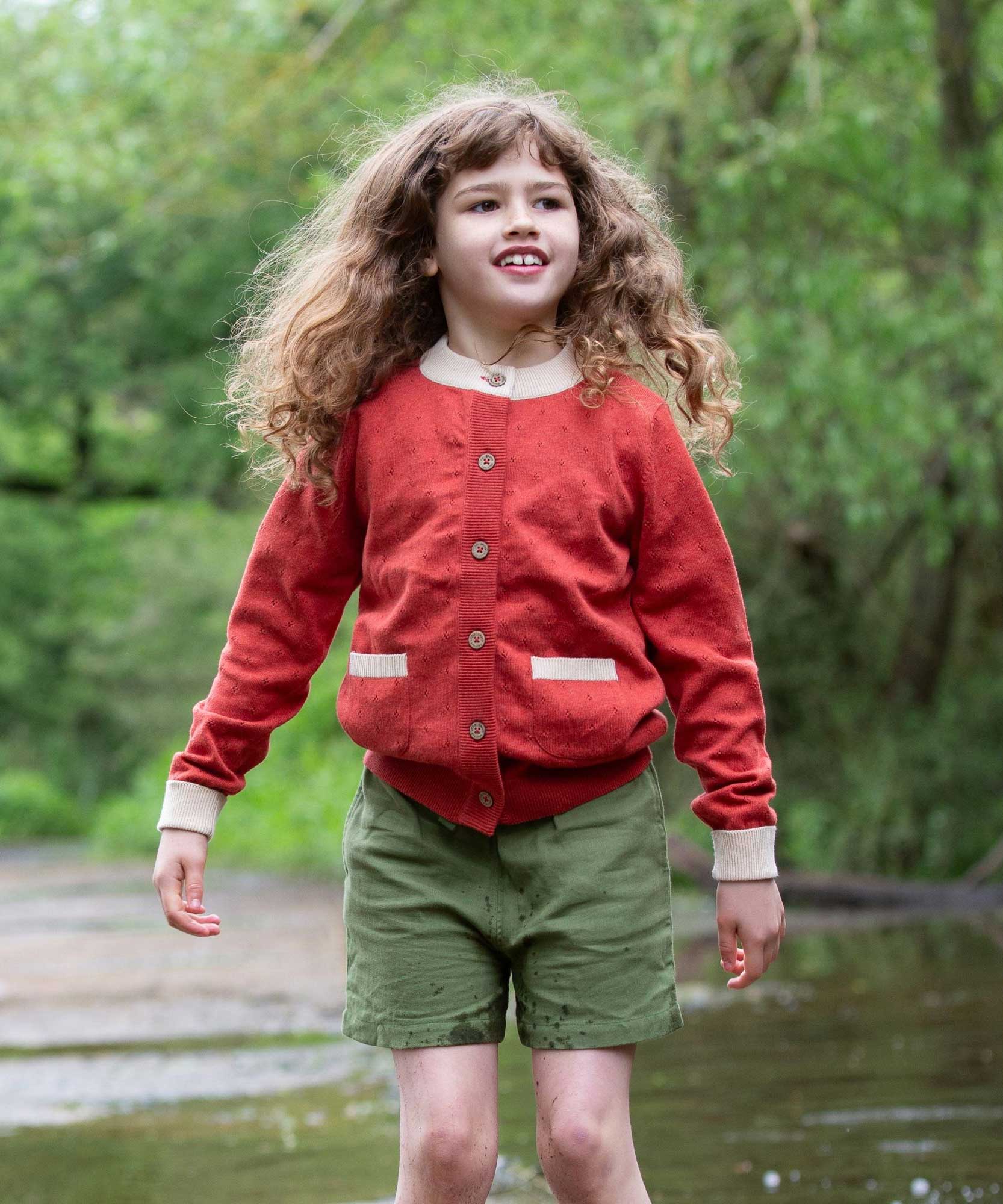A child wearing a Little Green Radicals From One To Another Burnt Ochre Pointelle Knitted Cardigan with green coloured shorts. Little green Radicals have a wide range of organic cotton, Fairtrade children's clothing and outerwear available here at Babipur.