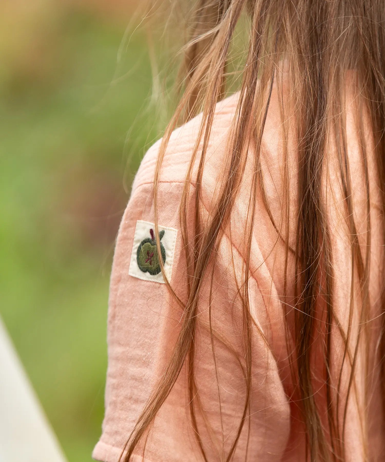Close up of the apple tag on the arm of the Little Green Radicals kids light coral pink short sleeved henley top being worn by a child.