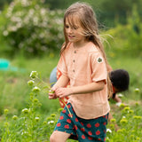 Child wearing the Little Green Radicals kids light coral pink short sleeved henley top with apple patch label on the side of one arms and a button up front with repeat red apple print dark blue shorts. 