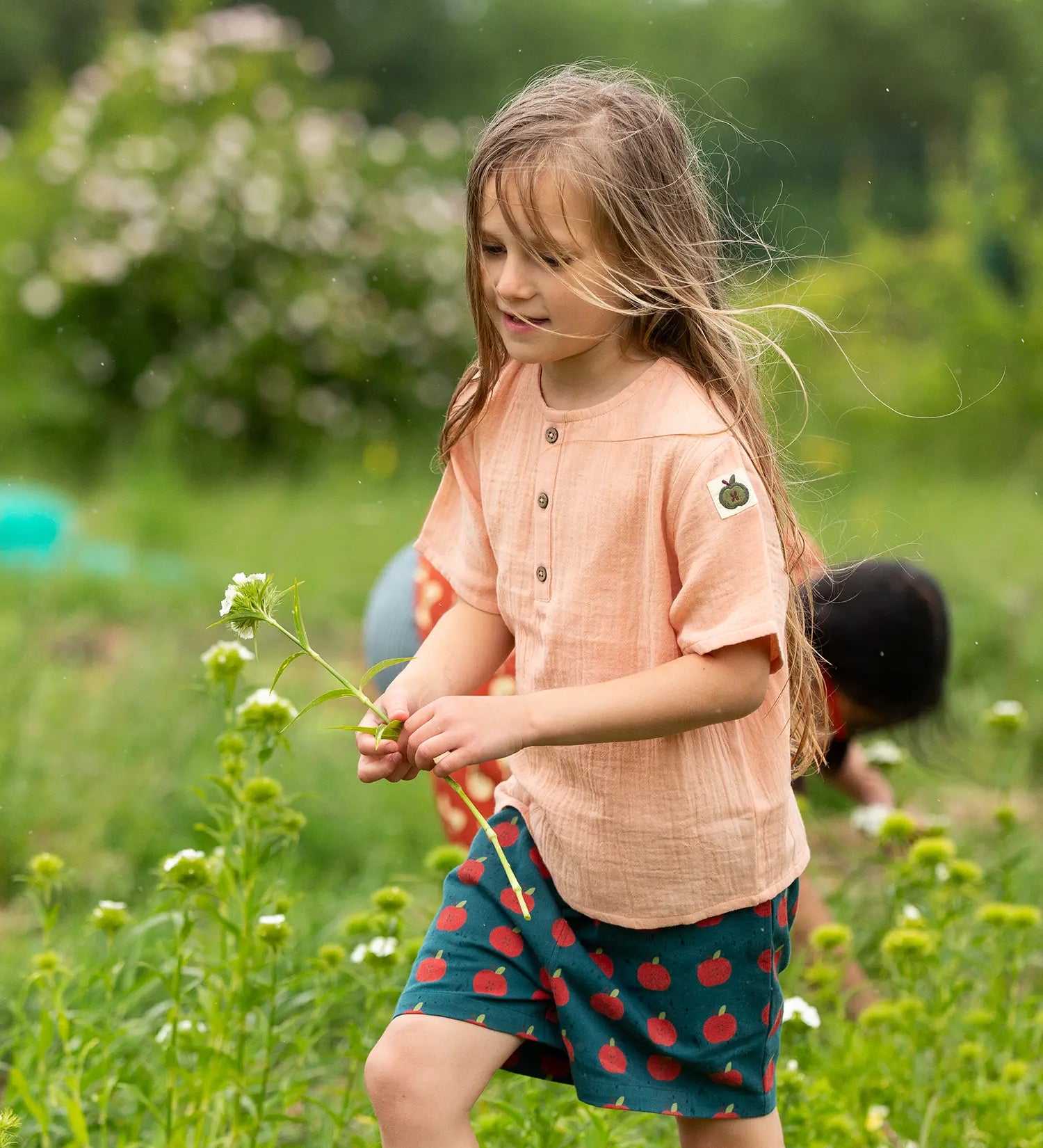 Child wearing the Little Green Radicals kids light coral pink short sleeved henley top with apple patch label on the side of one arms and a button up front with repeat red apple print dark blue shorts. 