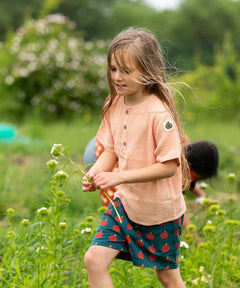 Child wearing the Little Green Radicals kids light coral pink short sleeved henley top with apple patch label on the side of one arms and a button up front with repeat red apple print dark blue shorts. 