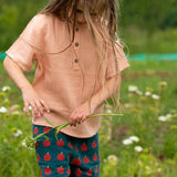 Child wearing the Little Green Radicals kids light coral pink short sleeved henley top with apple patch label on the side of one arms and a button up front with repeat red apple print dark blue shorts. 
