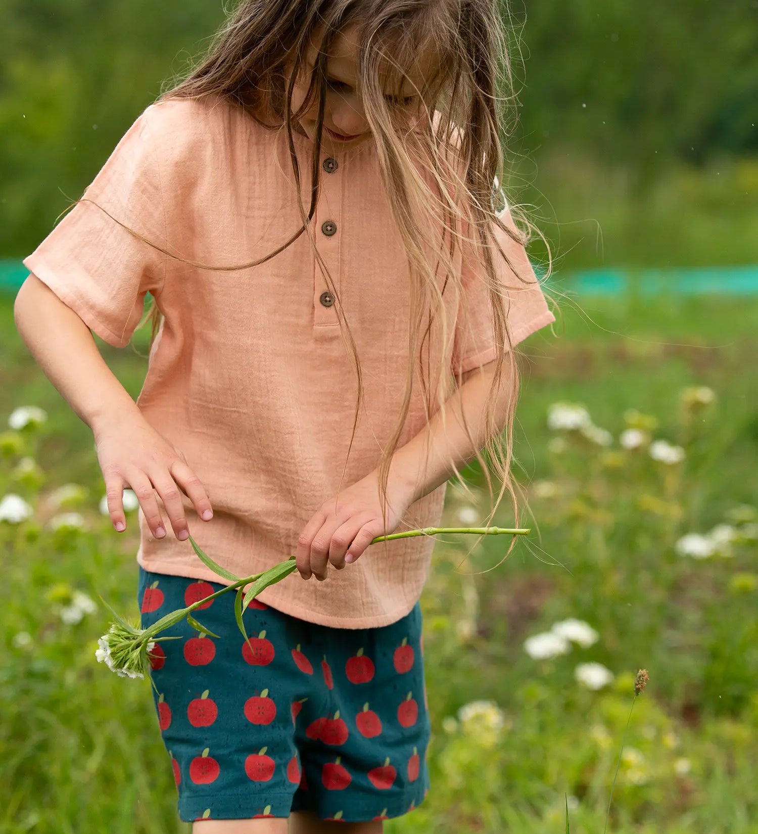 Child wearing the Little Green Radicals kids light coral pink short sleeved henley top with apple patch label on the side of one arms and a button up front with repeat red apple print dark blue shorts. 