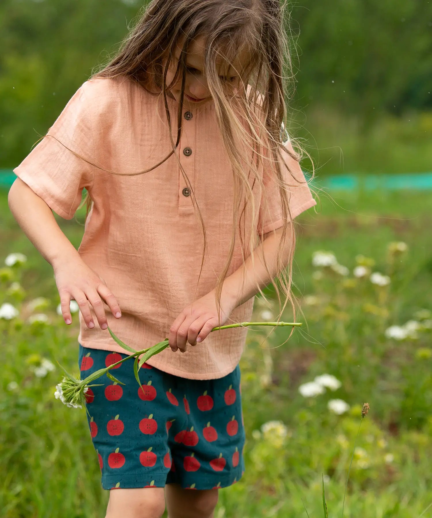 Child wearing the Little Green Radicals kids light coral pink short sleeved henley top with apple patch label on the side of one arms and a button up front with repeat red apple print dark blue shorts. 