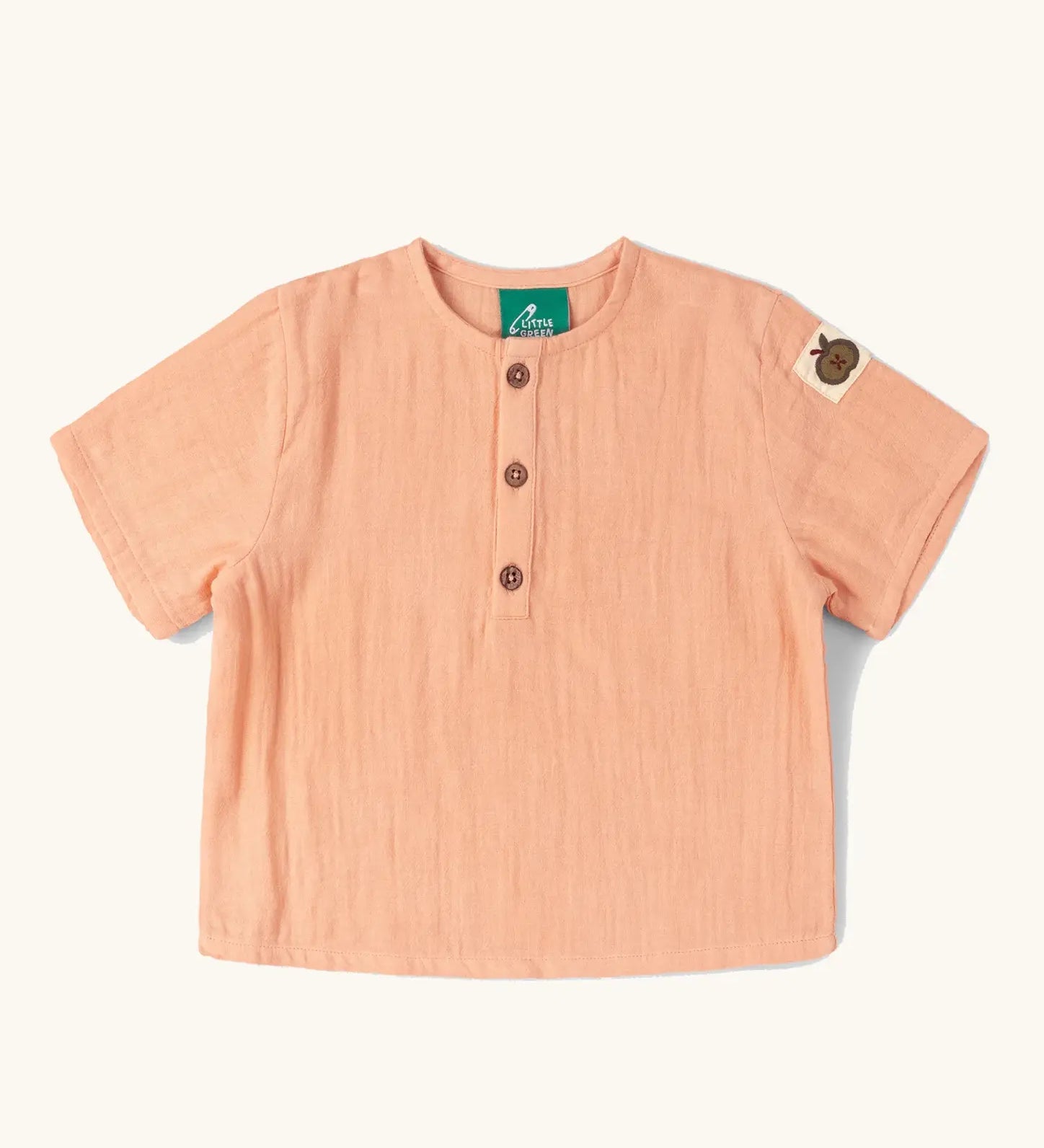 Little Green Radicals kids light coral pink short sleeved henley top with apple patch label on the side of one arms and a button up front.