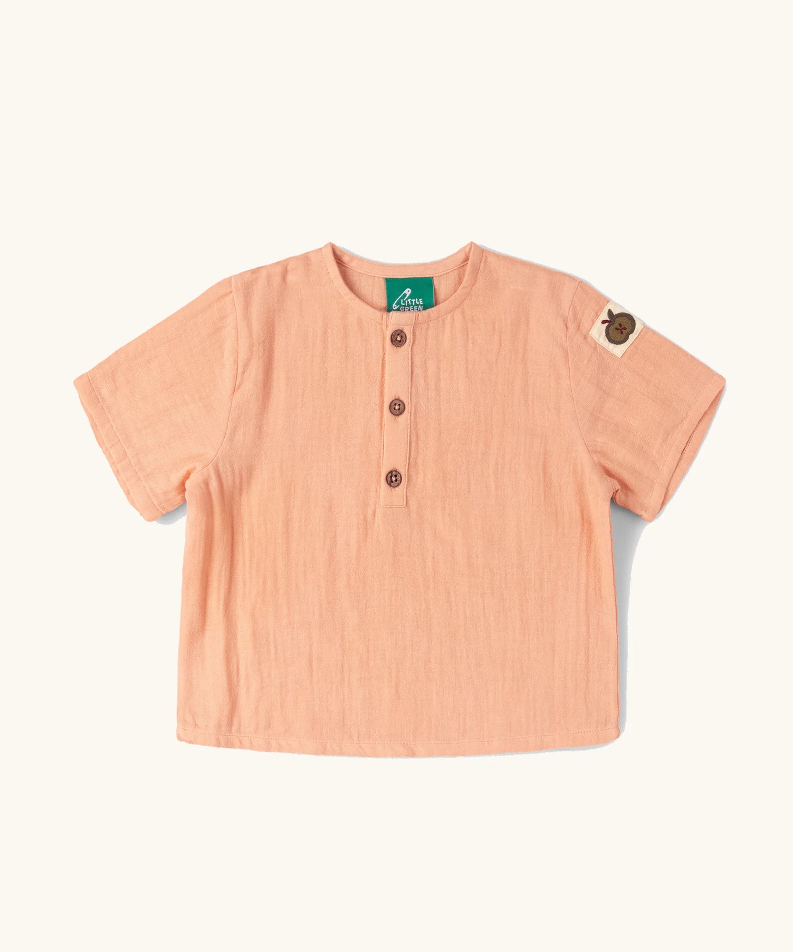 Little Green Radicals kids light coral pink short sleeved henley top with apple patch label on the side of one arms and a button up front.