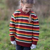 Child wearing the Little Green Radicals knitted snuggly jumper in a  hazel rainbow striped design with blue corduroy trousers. 