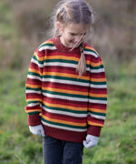 Child wearing the Little Green Radicals knitted snuggly jumper in a  hazel rainbow striped design with blue corduroy trousers. 