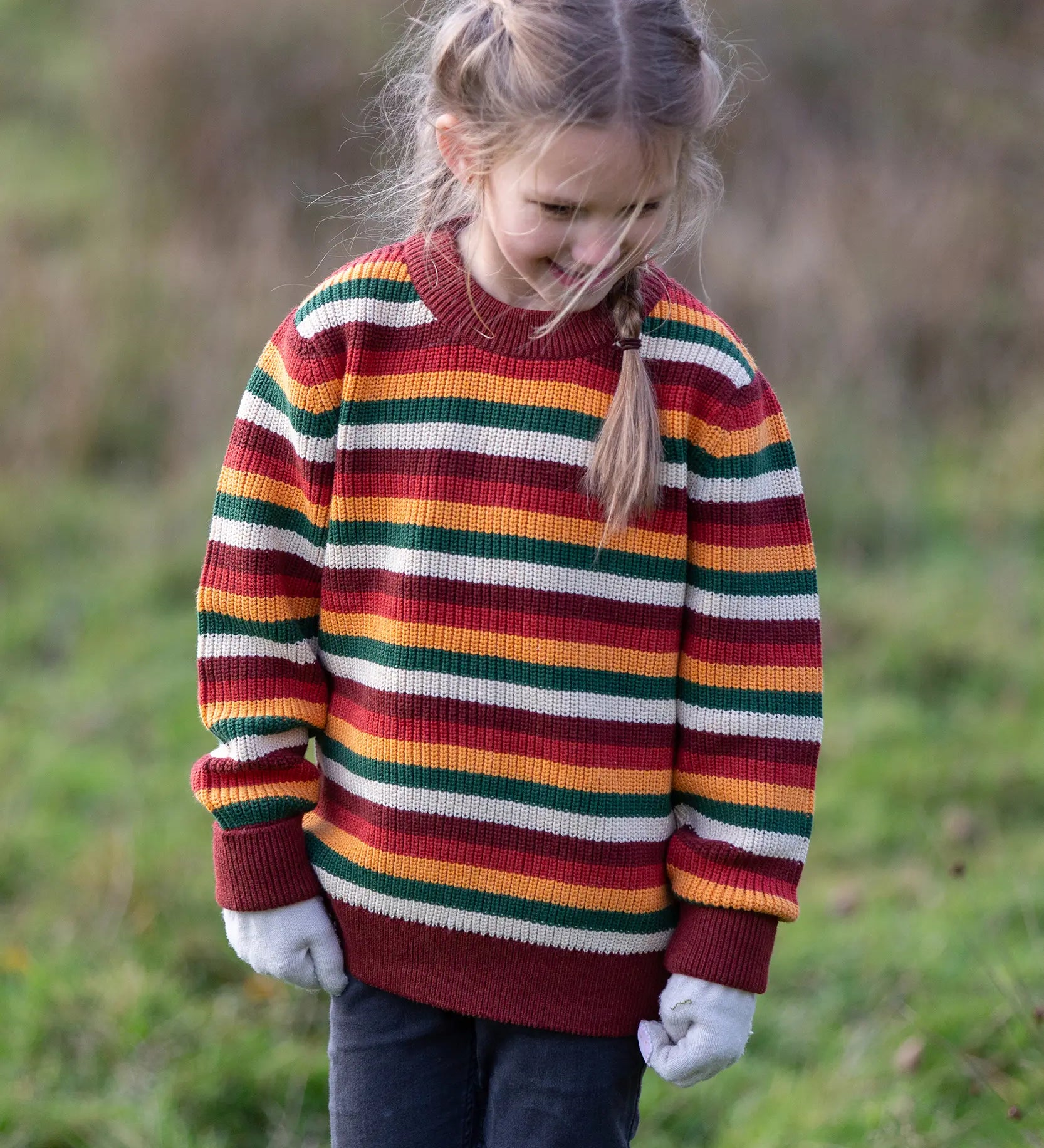 Child wearing the Little Green Radicals knitted snuggly jumper in a  hazel rainbow striped design with blue corduroy trousers. 