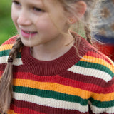 Close up of a child wearing the Little Green Radicals knitted snuggly jumper in a hazel rainbow striped design.