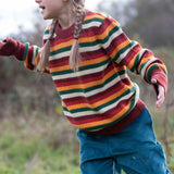 Child wearing the Little Green Radicals knitted snuggly jumper in a  hazel rainbow striped design  with blue corduroy trousers. 