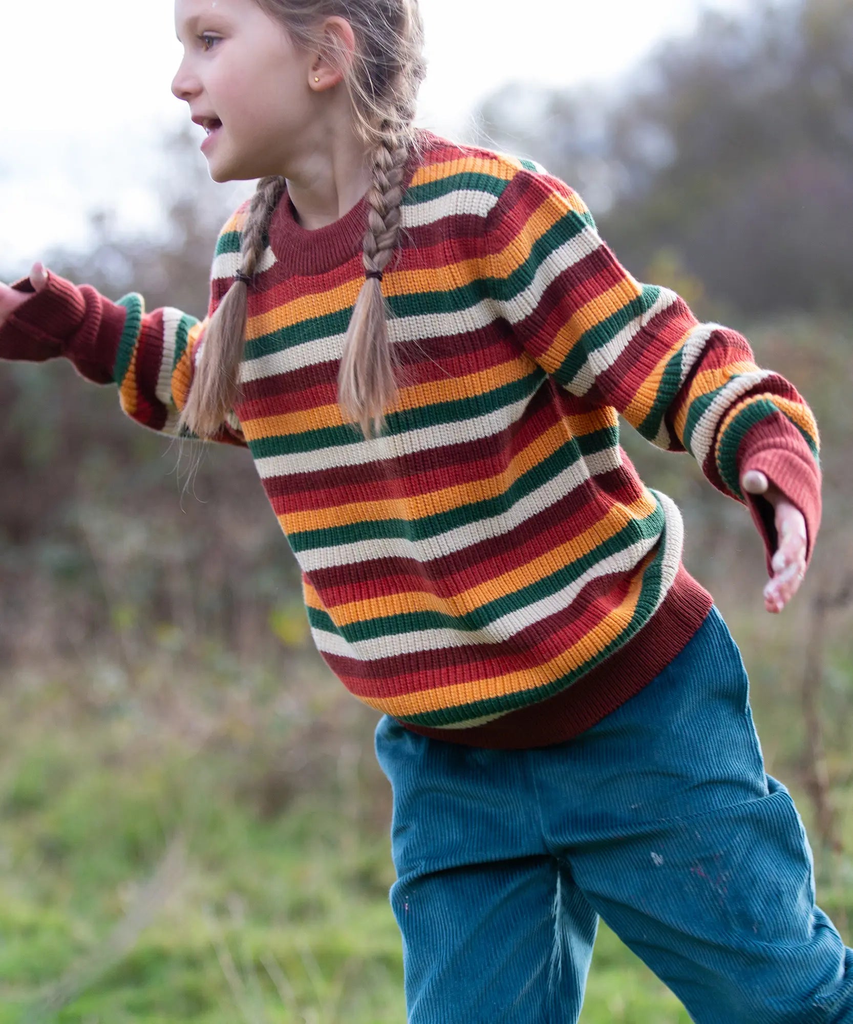 Child wearing the Little Green Radicals knitted snuggly jumper in a  hazel rainbow striped design  with blue corduroy trousers. 