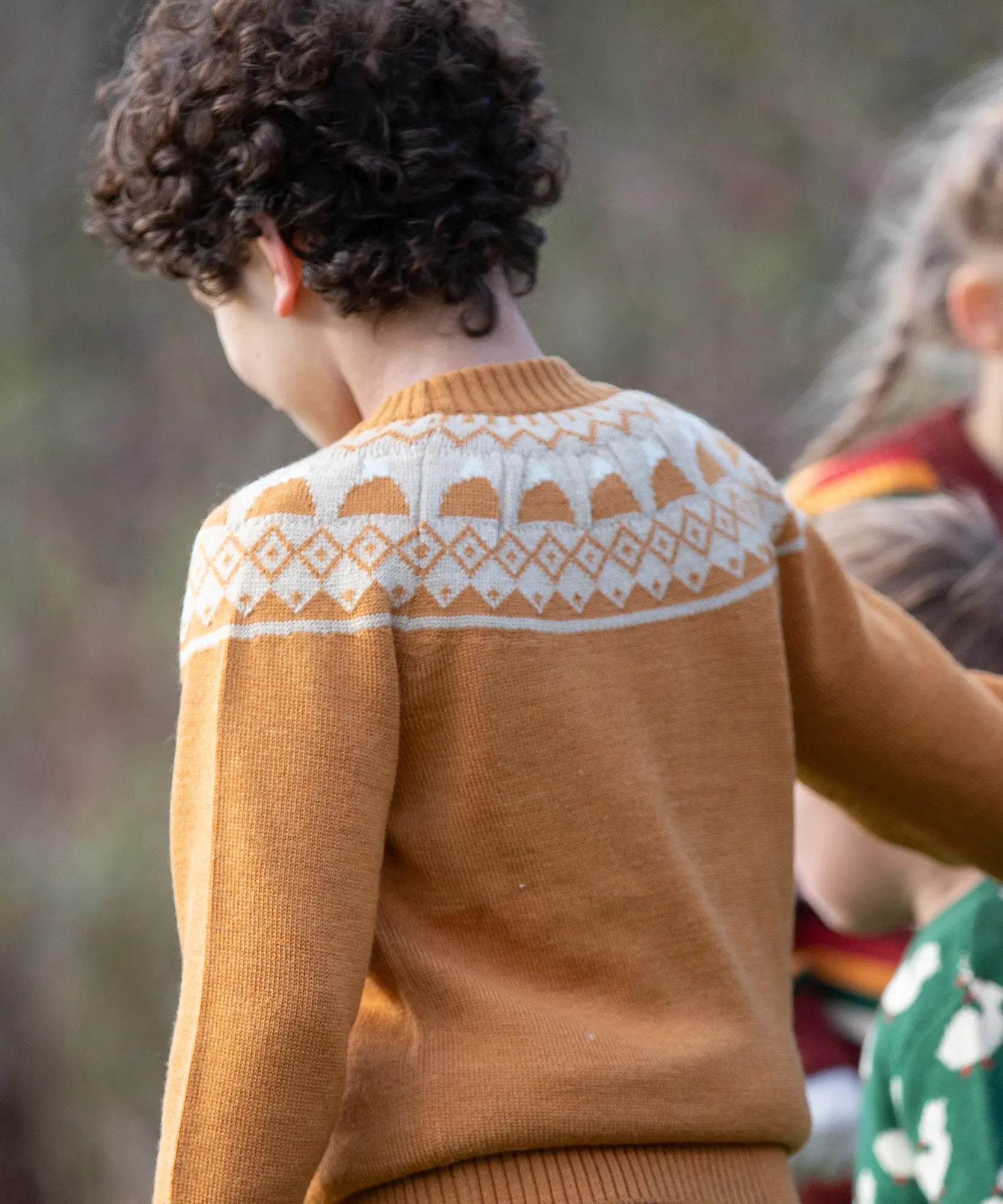 Back of a child wearing the Little Green Radicals mountain Fair Isle snuggly woollen jumper in yellow gold. The Fair Isle details are around the neckline featuring snow topped mountains surrounded by patterns.