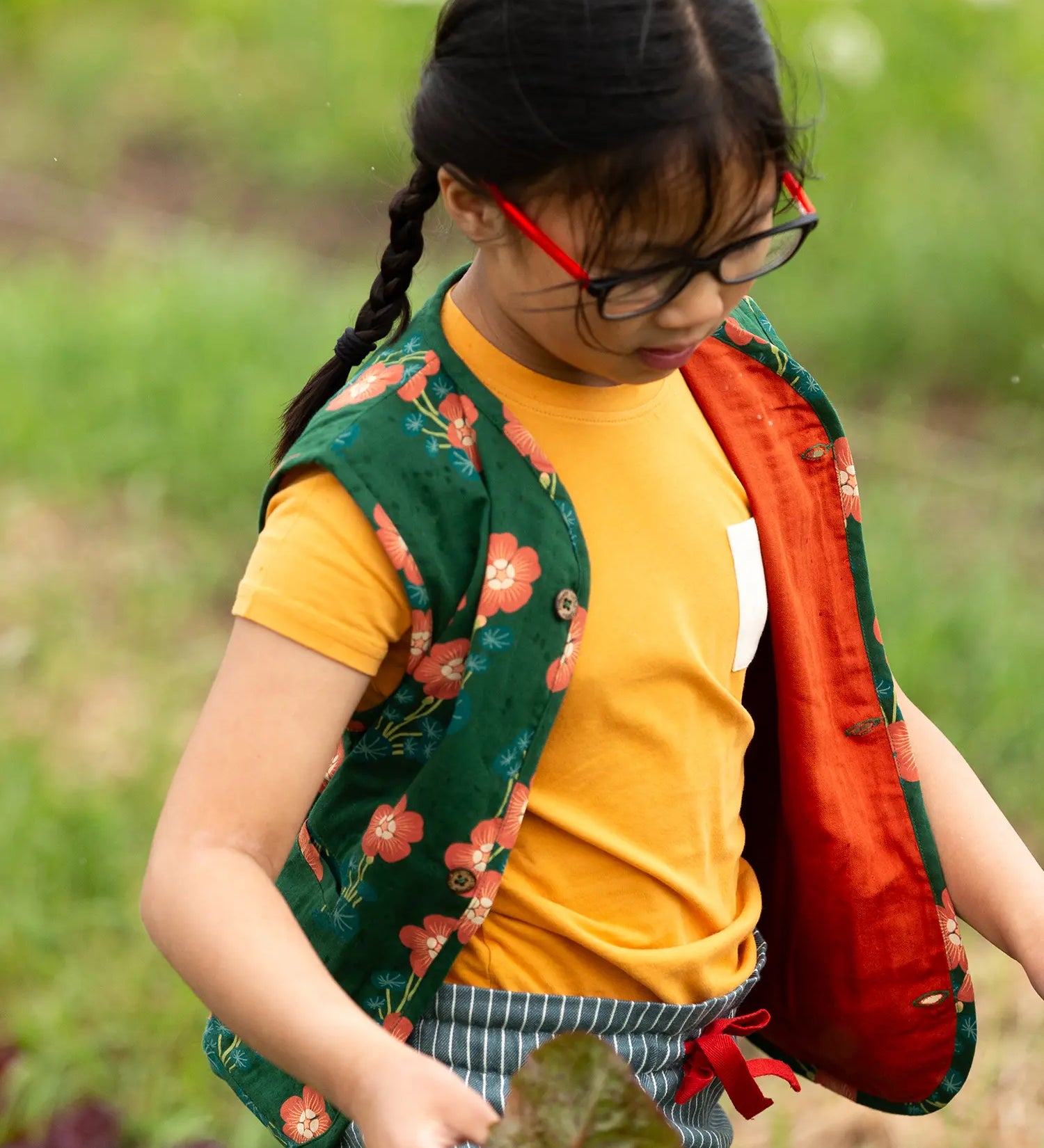 Child wearing a Little Green Radicals golden ochre yellow short sleeve essential short sleeve, organic cotton t-shirt underneath a green floral print gilet. 
 