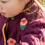 Close up of a child wearing the Little Green Radicals organic cotton snuggly knitted cardigan with a berry petal design. Dark purple cardigan with pink flowers details.