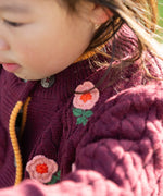 Close up of a child wearing the Little Green Radicals organic cotton snuggly knitted cardigan with a berry petal design. Dark purple cardigan with pink flowers details.
