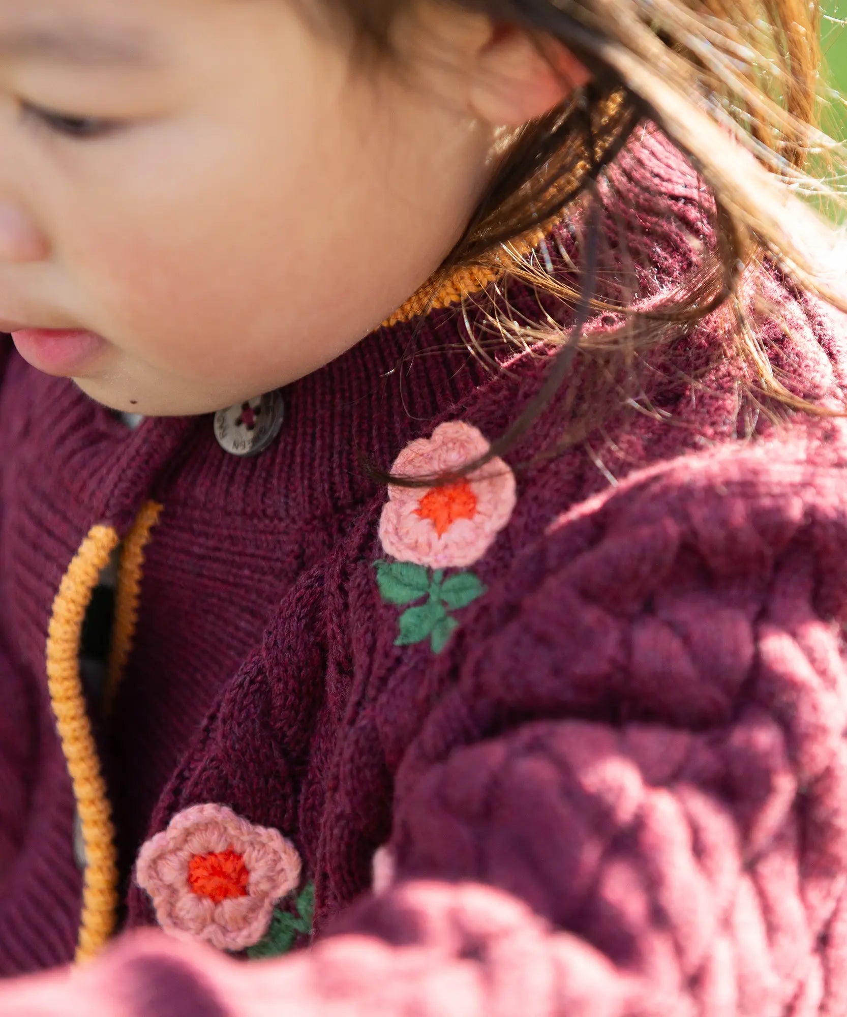 Close up of a child wearing the Little Green Radicals organic cotton snuggly knitted cardigan with a berry petal design. Dark purple cardigan with pink flowers details.