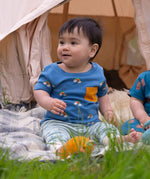 A young child sitting outdoors wearing the Little Green Radicals Kids' Organic T-shirt and Joggers Playset outfit in the  Blue Rainbows design. 